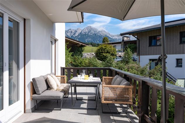 A cozy terrace with a table and chairs, surrounded by a beautiful mountain landscape. The sun is shining and the sky is blue.