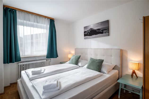 A cozy bedroom with a large bed and two pillows. The walls are light, and the window is decorated with curtains in blue and white.