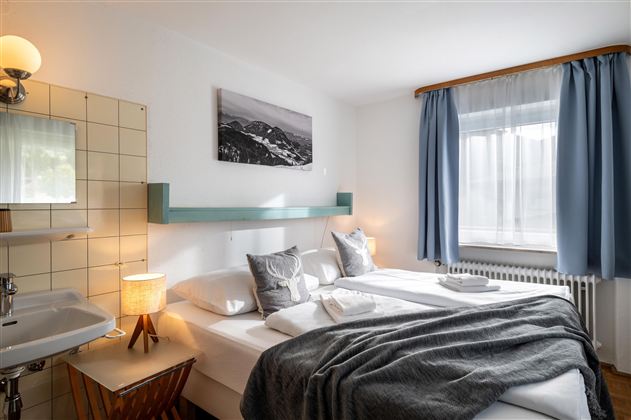 A cozy bedroom with a double bed and two pillows. The room has large windows and a calm, friendly atmosphere.