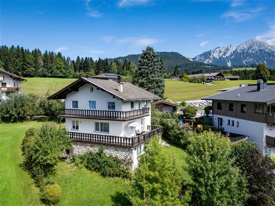 A beautiful house in a rural area, surrounded by green fields and trees. In the background, mountains and a blue sky can be seen.