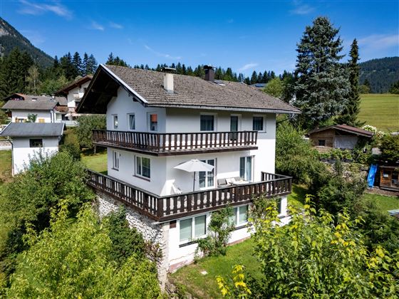 A cozy house with several balconies, surrounded by trees and meadows. In the background, mountains and a clear blue sky can be seen.