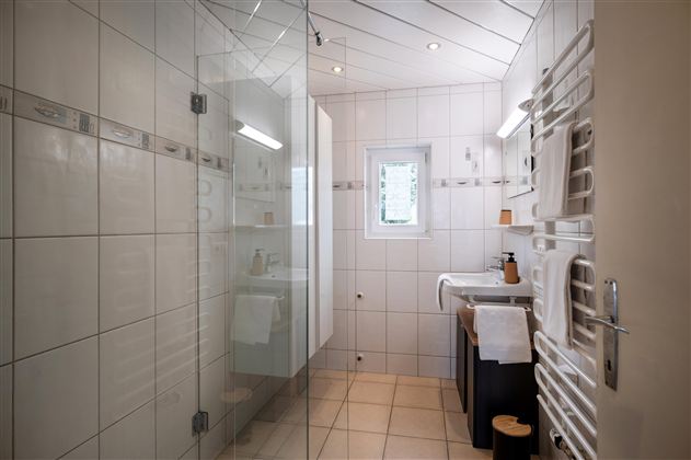 A modern bathroom with a glass shower and bright tiles. On the wall, there is a towel warmer, and a large window provides daylight.