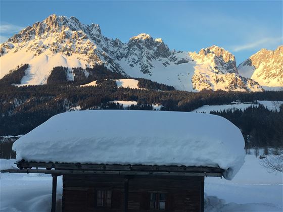 A cozy wooden house with a snow-covered roof. In the background, majestic mountains rise under a clear sky.