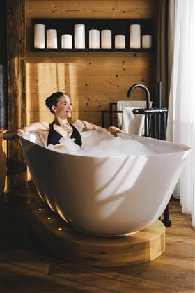 Eine Frau entspannt in einer modernen Badewanne mit Schaum. Der Raum hat warmes Holz und sanfte Beleuchtung.