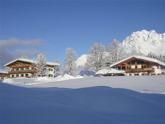 A beautiful winter landscape with snow-covered huts and tall mountains in the background. The clear blue sky completes the idyllic scene.