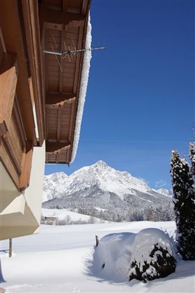 Eine Winterlandschaft mit schneebedeckten Hügeln und Bergen unter einem klaren blauen Himmel. Im Vordergrund ist ein Teil eines Hauses zu sehen.