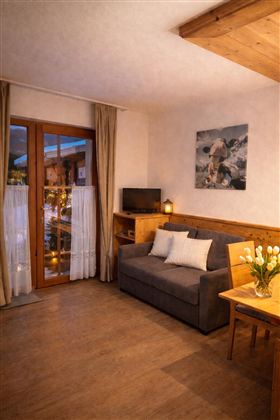 A cozy living room with a gray sofa and a wooden table. Large windows with curtains provide plenty of daylight and a warm atmosphere.