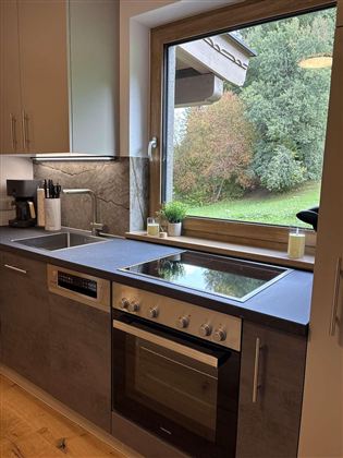 Eine moderne Küche mit einem großen Fenster, das auf einen grünen Garten blickt. Auf der Arbeitsplatte befinden sich Herd und Spüle.