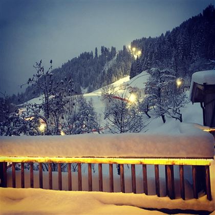Een besneeuwd landschap met verlichte bomen en bergen op de achtergrond. De lucht is grijs en schemerig, wat een rustige winterstemming creëert.