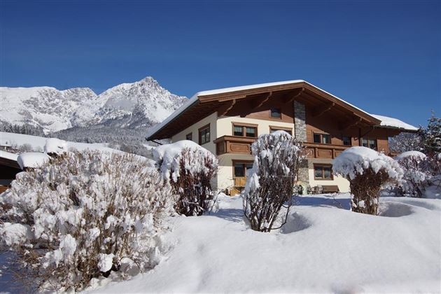 Ein wunderschönes, schneebedecktes Haus vor einer malerischen Bergkulisse. Die Umgebung ist ruhig und winterlich.