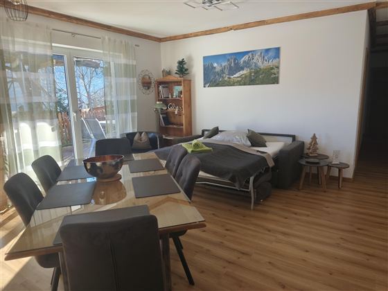 Een lichte en uitnodigende woonkamer met een eettafel en stoelen. Op de achtergrond is een gezellig bed en een raam met uitzicht op de natuur.