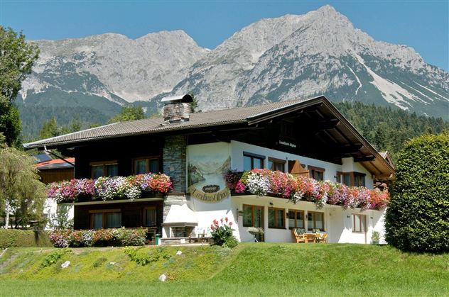 Ein malerisches Haus mit bunten Blumenfenstern in einer grünen Umgebung. Im Hintergrund erhebt sich ein beeindruckendes Gebirge unter klarem Himmel.