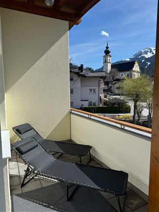 A balcony with two loungers and a beautiful view of a small village. In the background, the mountains and a church tower are visible.