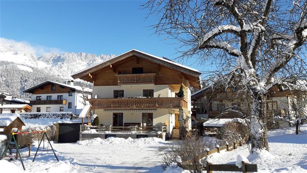 Ein gemütliches Haus im Schnee mit einem klaren blauen Himmel. Umgeben von Bergen und anderen Gebäuden, bietet es eine idyllische Winterlandschaft.