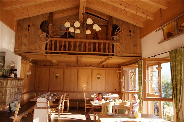 Een gezellige kamer met houten wandbekleding en grote ramen. Boven is een balkon zichtbaar, en de tafels zijn vriendelijk gedekt.