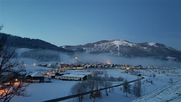 Eine verschneite Landschaft im Morgengrauen mit sanften Hügeln und einem kleinen Dorf. Die Lichter der Häuser leuchten sanft durch den Nebel.