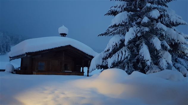 Eine gemütliche Berghütte im Schnee unter einem grauen Himmel. Der Boden ist mit einer dicken Schneeschicht bedeckt.