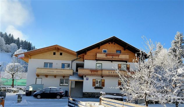 Ein gemütliches Haus im Schnee mit Holzverzierungen. Die Umgebung ist winterlich und von Bäumen umgeben.