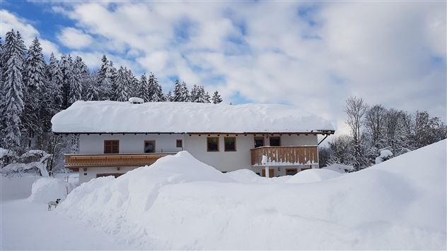 Ein schneebedecktes Haus umgeben von hohen Schneebergen. Der Himmel ist klar mit einigen Wolken.