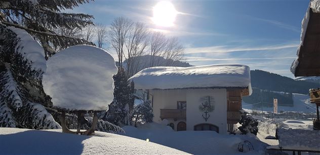 Eine verschneite Landschaft mit einem häuslichen Gebäude und einem großen, schneebedeckten Baum im Vordergrund. Die Sonne scheint hell am blauen Himmel.