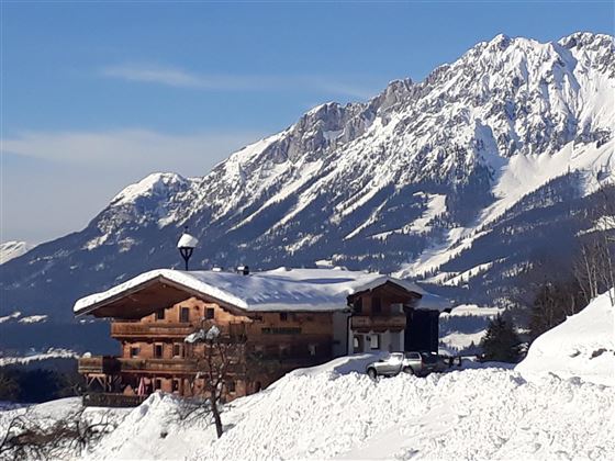 A charming wooden house in the snow with impressive mountains in the background. The landscape is wintery and peaceful.