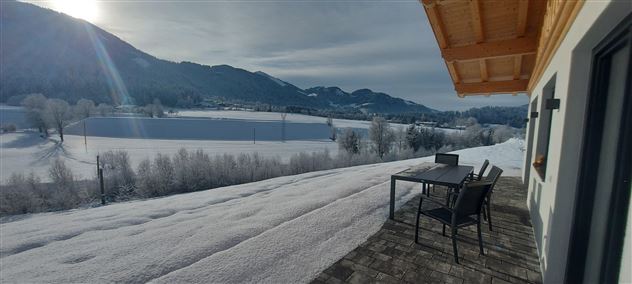 Eine schneebedeckte Landschaft mit Bergen im Hintergrund. Auf der Terrasse stehen ein Tisch und Stühle, ideal zum Entspannen.