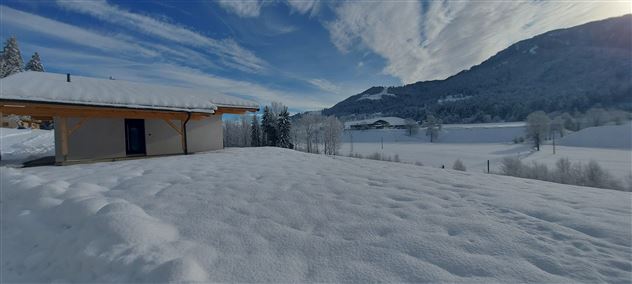 Eine schneebedeckte Landschaft mit einem Haus im Vordergrund. Im Hintergrund sind Berge und ein blauer Himmel mit Wolken sichtbar.
