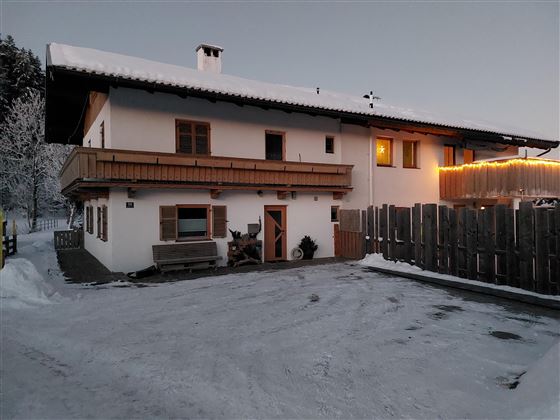 Ein gemütliches Haus im Schnee mit einem Balkons und warmem Licht. Der Parkplatz ist schneebedeckt und es gibt einen Holzzaun.