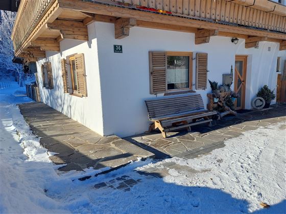 Ein gemütliches Haus im Schnee mit Holzfensterläden und einer Bank vor der Tür. Der Weg ist mit Steinen belegt und das Umfeld ist winterlich.