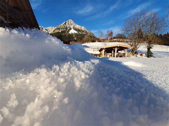 A snowy landscape with a view of a mountain in the background. In the foreground lies a slope with fresh snow, next to it is a cozy cabin.