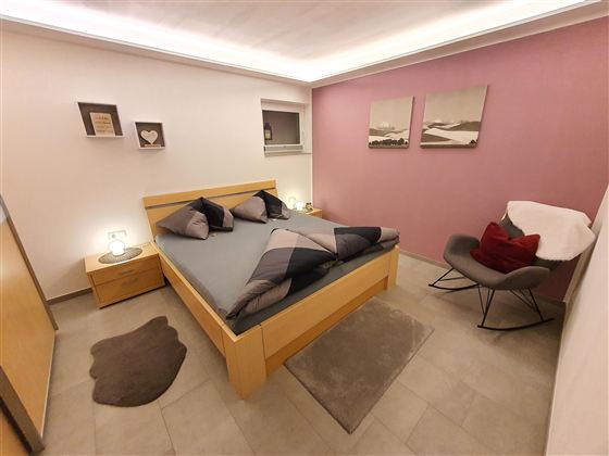 Ein modernes Schlafzimmer mit einem Holzbett und gemütlichen Kissen. Die Wände sind in sanften Farben gehalten, und ein bequemer Sessel steht in der Ecke.