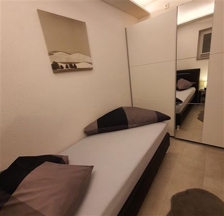 Ein einfaches Schlafzimmer mit einem Einzelbett, großen Kissen und einem Spiegel. An der Wand hängt ein Bild von einer Landschaft.