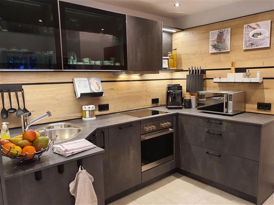 Eine moderne Küche mit grauen Möbeln und Holzdetails. Sie ist mit verschiedenen Geräten wie einem Herd, einer Mikrowelle und einem Spülbecken ausgestattet.