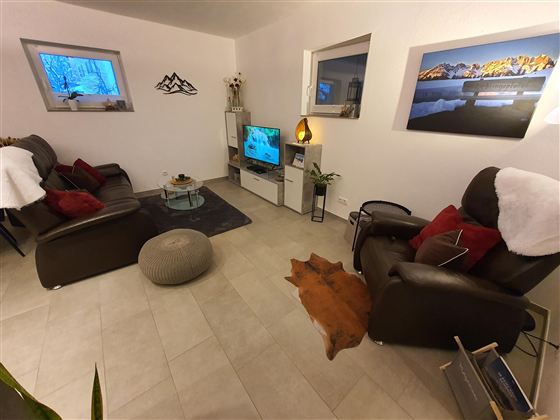 Ein gemütliches Wohnzimmer mit Fernseher, zwei bequemen Sesseln und einer Couch. Die Wände sind mit Bildern dekoriert und es gibt Böden aus hellen Fliesen.