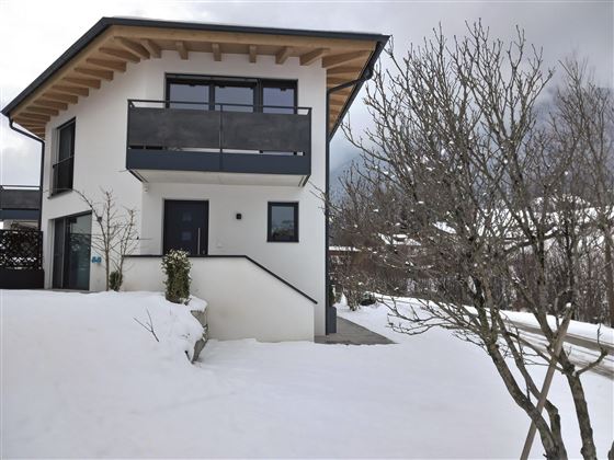 A modern house with a large balcony, surrounded by snow. In the background, bare trees are visible.