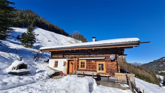 An idyllic mountain cabin in the snow, surrounded by forests and mountains. The blue sky and the snowy landscape create a relaxed winter atmosphere.