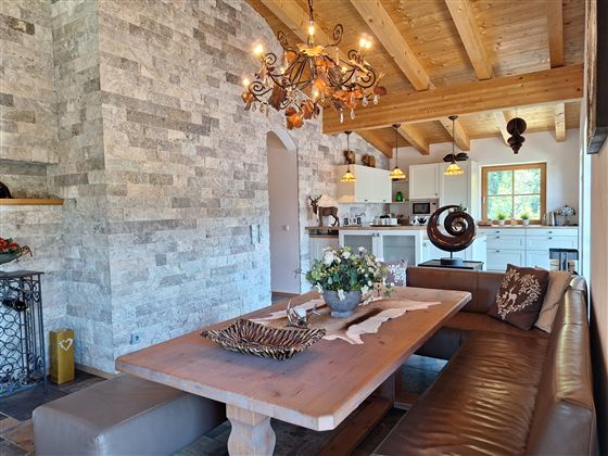 Een gezellige eetruimte met een grote houten tafel en zitplaatsen. De muren zijn bekleed met natuursteen, en er is een lichte, uitnodigende keuken op de achtergrond.