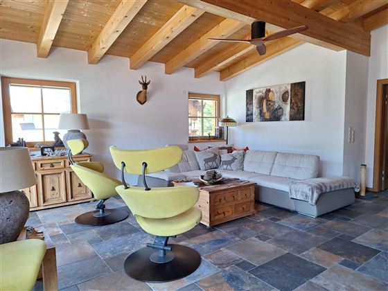 Een moderne woonkamer met houten plafonds en tegelvloeren. Er zijn een lichte bank, opvallende gele stoelen en decoratieve elementen te zien.