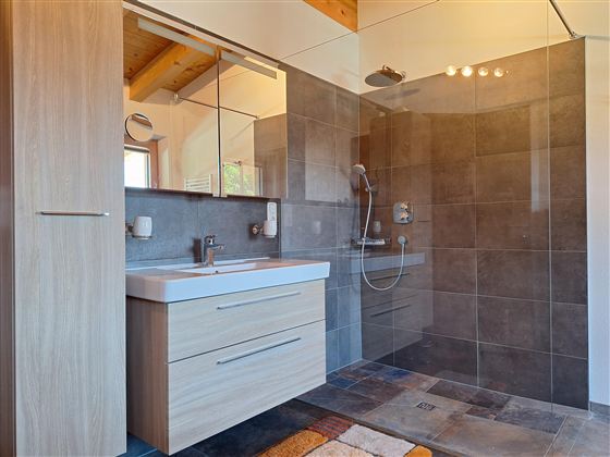 Een moderne badkamer met een ruime douche en houten details. Het wasbakgebied heeft een lichte houten commode en grote spiegels.