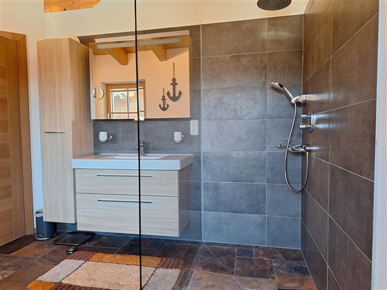 Een moderne badkamer met een douche en een wastafel in houtlook. De muren zijn voorzien van grijze tegels en er is een spiegel.