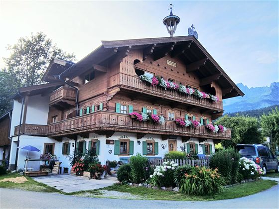 A traditional alpine house with wooden cladding and many flower balconies. The surroundings are green and sunny, with mountains in the background.