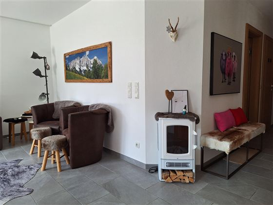 Een gezellige woonkamer met moderne meubels en een houtkachel. Aan de muur hangt een schilderij van bergen, en er is een kleine zithoek met kussens.