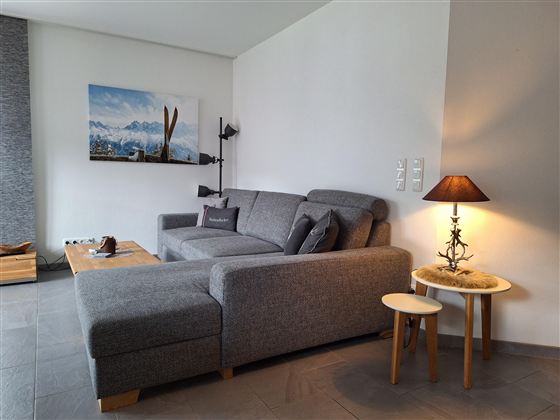 Ein gemütliches Wohnzimmer mit einem grauen Sofa und einem kleinen Beistelltisch. An der Wand hängt ein Bild von Berglandschaft mit Skiern.