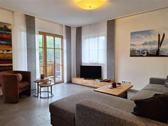 Ein modernes Wohnzimmer mit einer grauen Couch und einem kleinen Tisch. Große Fenster lassen viel Licht herein und bieten Zugang zu einem Balkon.