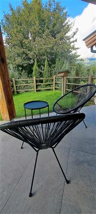 Twee moderne stoelen en een kleine tafel staan op een terras. Op de achtergrond zijn groene bomen en een hek te zien.