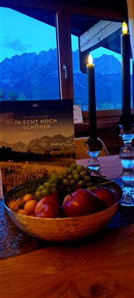 Een mooie tafel met een fruitschaal die appels en druiven bevat. Op de achtergrond staat een kaarsenhouder en een landschap foto voor een bergpanorama.