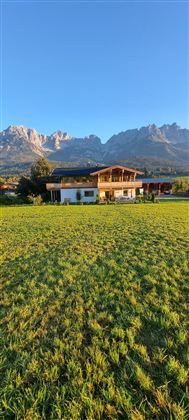 Een schilderachtig huis op een weiland met de bergen op de achtergrond. De lucht is helder en blauw.