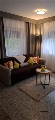 A cozy living room with a sofa and colorful cushions. The windows are covered with light curtains, and a small table stands in front of it.