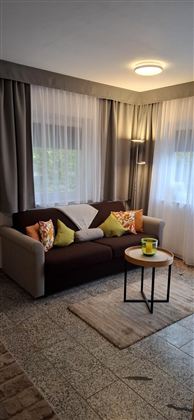 A cozy living room with a beige sofa and colorful cushions. There is a small table with a cup and large windows with curtains.
