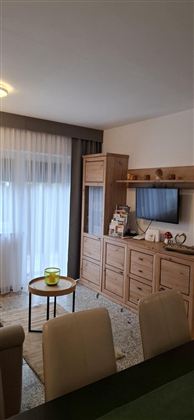 A cozy living space with wooden furniture and a television. Bright light streams through the curtains, creating an inviting atmosphere.